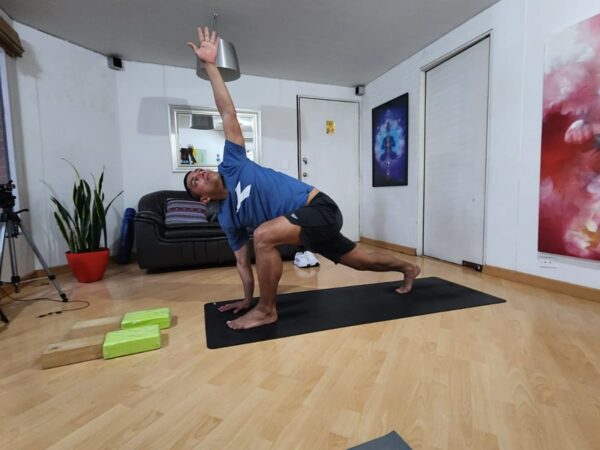 Trazando la Armonía: Exploraciones en Yoga Presencial Hombre en clase de yoga presencial, vistiendo camiseta azul y pantalones cortos negros, realiza una postura de zancada en esterilla negra. Su brazo derecho se alza mientras su mano izquierda toca el suelo. Bloques de yoga verdes y una toalla blanca están dispuestos cerca